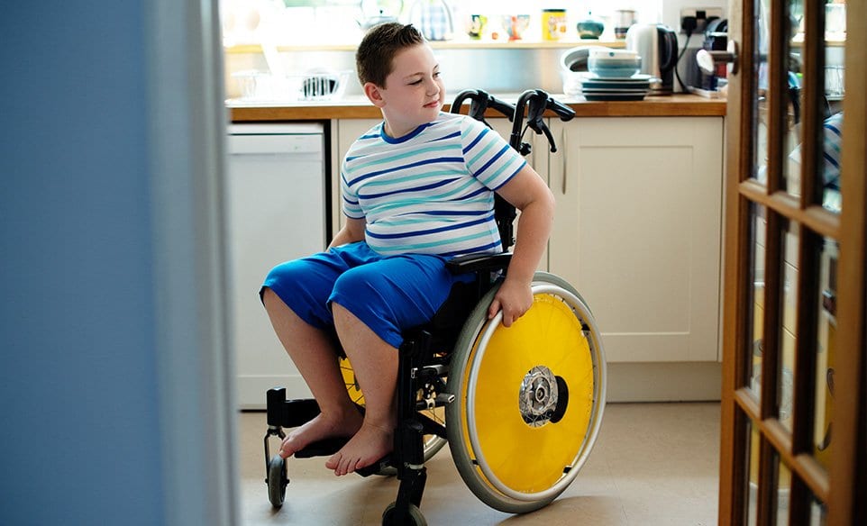 Young person in wheelchair, wearing stripey blue t shirt and blue shorts in a kitchen